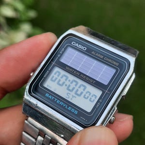 May include: A silver Casio digital watch with a black face displaying the time 00:00:00. The watch features the text "CASIO", "ALARM CHRONO", "WATER RESIST", and "BATTERYLESS". The watch band is silver.