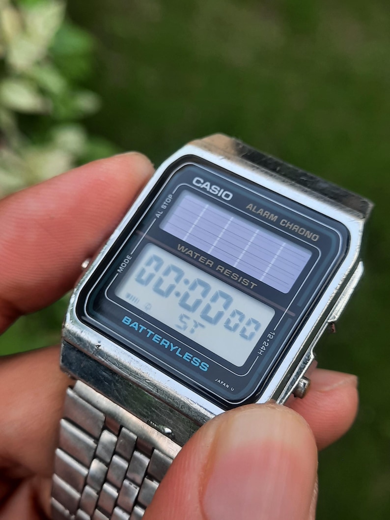 May include: A silver Casio digital watch with a black face displaying the time 00:00:00. The watch features the text "CASIO", "ALARM CHRONO", "WATER RESIST", and "BATTERYLESS". The watch band is silver.