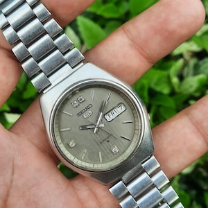 May include: A silver-toned Seiko automatic wristwatch with a gray dial. The watch features a day-date display, diamond-like markers, and a stainless steel band. The watch is shown against a green leafy background.