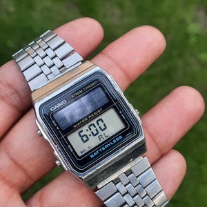 May include: A silver Casio digital watch displaying 6:00 on the screen. The watch has a black face with the words "WATER RESIST" and "BATTERYLESS". The watch band is made of silver metal links.