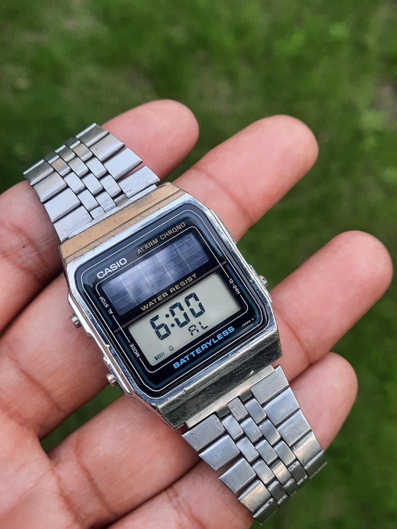 May include: A silver Casio digital watch displaying 6:00 on the screen. The watch has a black face with the words "WATER RESIST" and "BATTERYLESS". The watch band is made of silver metal links.