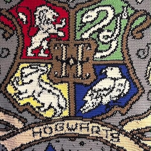 Può includere: Una coperta all'uncinetto grigia che presenta lo stemma di Hogwarts con una lettera H centrale, circondata da quadranti rossi, verdi, gialli e blu contenenti un leone, un serpente, un tasso e un'aquila. Sotto lo stemma c'è uno stendardo con il testo Hogwarts.