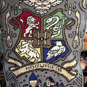 May include: A gray crocheted blanket featuring the Hogwarts crest. The crest is divided into four sections with a lion, snake, badger, and eagle. The word "Hogwarts" is on a banner below the crest. The blanket also includes images of a broom, candles, and an owl.