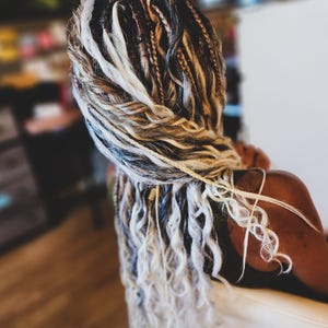 May include: Close-up of braided hair with a mix of colors, including blonde, black, and brown. The braids are styled in a layered pattern, with some sections tightly woven and others loose and wavy. The hair is styled in a way that is both intricate and visually appealing.