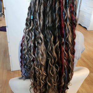May include: Long, curly hair in shades of brown, blonde, red, and blue, styled with dreadlocks and beads. The person is seated in a white chair. The hair has a natural, textured appearance.