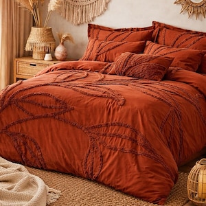 以下が含まれることがあります： 葉のようなテクスチャードデザインの、錆色の寝具セット。セットには、掛け布団カバーといくつかの枕が含まれています。ベッドは織りのラグの上にあり、部屋は装飾要素を備えたボヘミアンスタイルです。