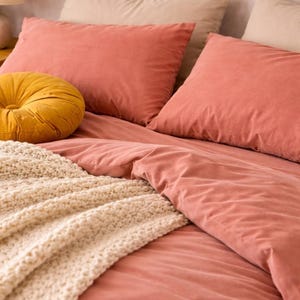 Op de afbeelding: Een bed met koraalkleurig beddengoed, inclusief kussens en een dekbed. Een crèmekleurige, grof gebreide deken is over het bed gedrapeerd. Een rond, mosterdgeel kussen voegt een contrasterend accent toe.