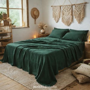 Op de afbeelding: Een bed is bedekt met een donkergroene linnen lakenset, inclusief een laken, hoeslaken en kussenslopen. Het bed staat op een lichtgekleurd tapijt. De kamer heeft een bohemien stijl met macramé wandkleden en een strohoed.