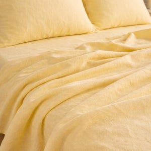 May include: A close-up shot of a bed with yellow bedding. The set includes two pillowcases and a fitted sheet. The fabric has a textured appearance, and the color is a soft, pale yellow. The bedding is neatly arranged, suggesting a comfortable and inviting bedroom setting.