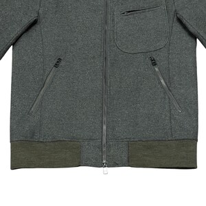 May include: A dark gray zip-up jacket with a small pocket on the chest and two zippered pockets on the front. The jacket has a ribbed, olive green hem and cuffs. The zipper is silver.