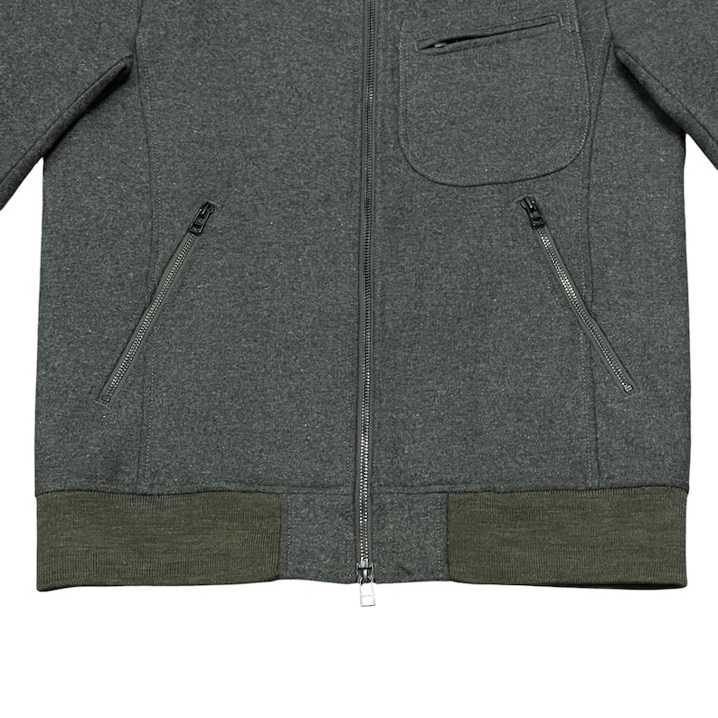 May include: A dark gray zip-up jacket with a small pocket on the chest and two zippered pockets on the front. The jacket has a ribbed, olive green hem and cuffs. The zipper is silver.