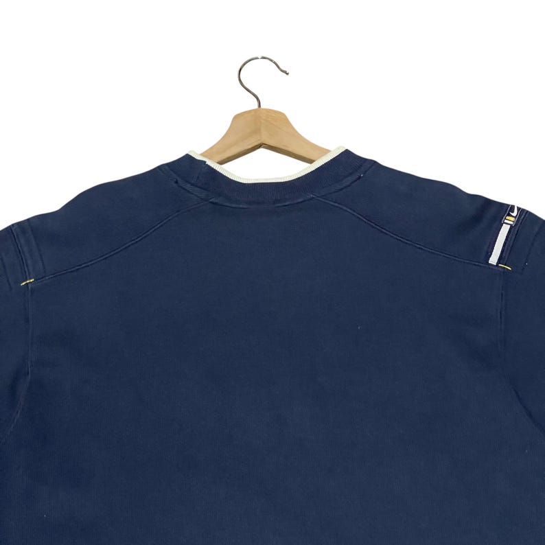 Puede incluir: Sudadera azul marino con cuello blanco y detalles de costura amarillos. La sudadera est&aacute; colgada de una percha de madera. El hombro tiene una raya blanca y amarilla.