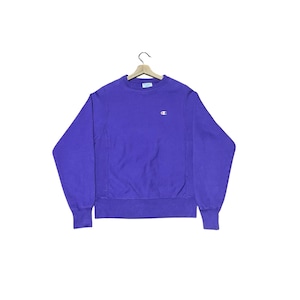 Puede incluir: Sudadera morada de cuello redondo Champion. La sudadera tiene un pequeño logotipo blanco en el pecho izquierdo. Cuelga de una percha de madera sobre un fondo blanco. Los puños y el dobladillo son acanalados.