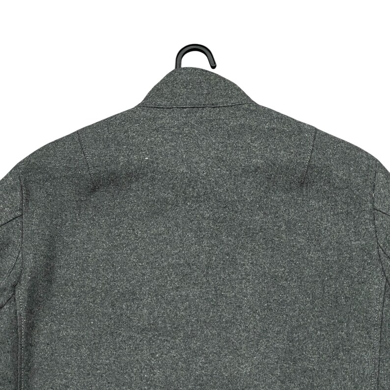 May include: A gray wool coat, viewed from the back, hanging on a black hanger. The coat has a classic collar and a tailored fit. The fabric has a textured appearance.