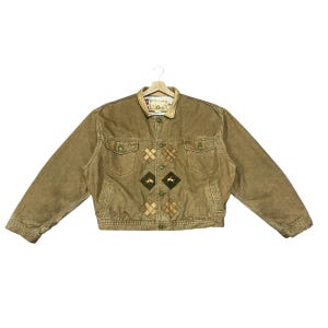 Puede incluir: Chaqueta vaquera marrón con cuello de pana y parches en forma de diamante. La chaqueta tiene dos bolsillos en el pecho y dos bolsillos laterales. Los parches presentan diseños bordados. La chaqueta está colgada de una percha de madera.