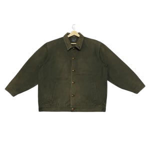 May include: An olive green jacket with a button-up front, two front pockets, and a collared neckline. The jacket is made of a durable fabric and has long sleeves. The jacket is hanging on a wooden hanger.