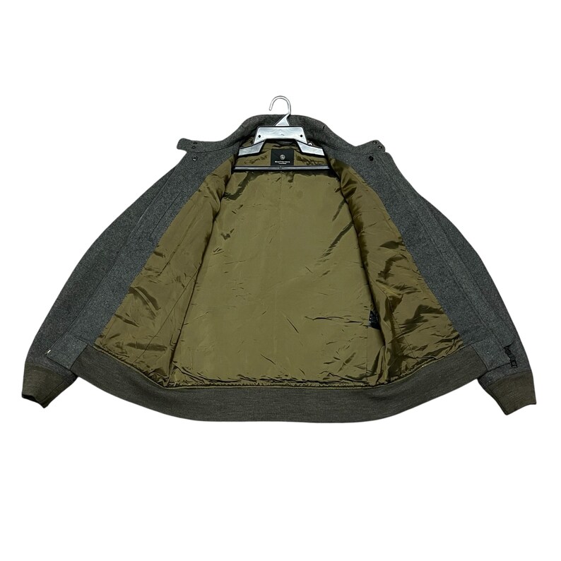 May include: A gray wool jacket with a dark olive green interior lining, hanging on a white hanger. The jacket has a button-up front and a ribbed hem and cuffs. The jacket is open, revealing the interior lining.