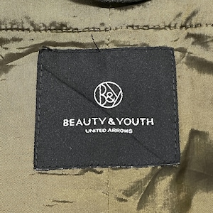 May include: A close-up of a black label with the text "BEAUTY & YOUTH UNITED ARROWS" in white. The label has a circular logo with the letters "B&Y" inside. The background is olive green.