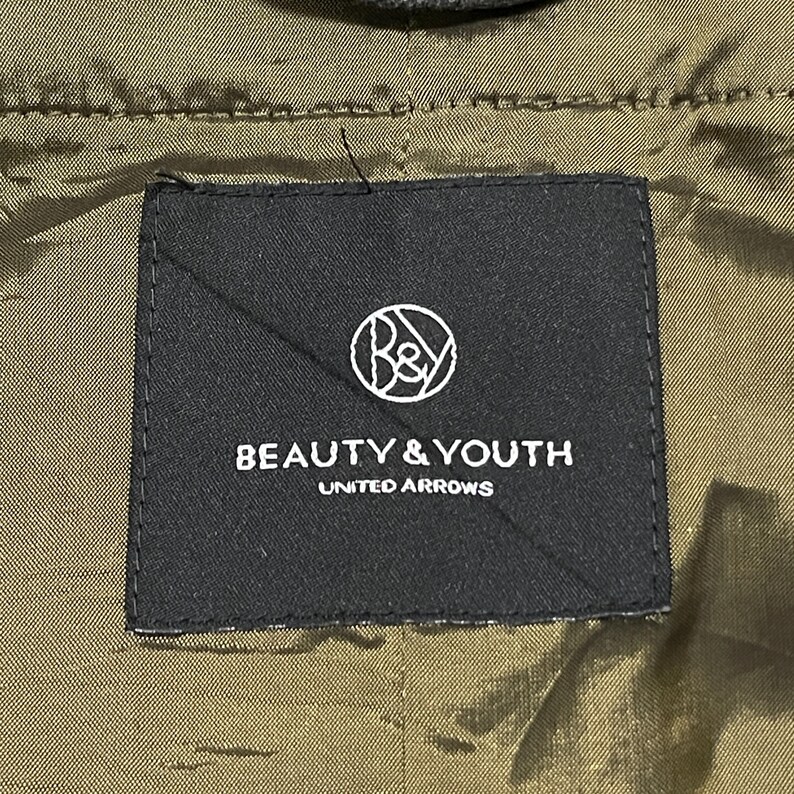 May include: A close-up of a black label with the text "BEAUTY & YOUTH UNITED ARROWS" in white. The label has a circular logo with the letters "B&Y" inside. The background is olive green.