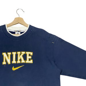 Puede incluir: Sudadera Nike azul marino con cuello blanco y azul marino. La palabra "NIKE" est&aacute; bordada en amarillo en el pecho, con un logotipo de Nike swoosh amarillo debajo. La sudadera est&aacute; colgada en una percha de madera.