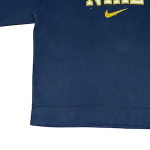 Puede incluir: Sudadera azul marino con la palabra "NIKE" en letras amarillas y un logotipo de swoosh amarillo. La sudadera tiene un dobladillo acanalado y cuello redondo. La tela parece ser un material suave y c&oacute;modo.