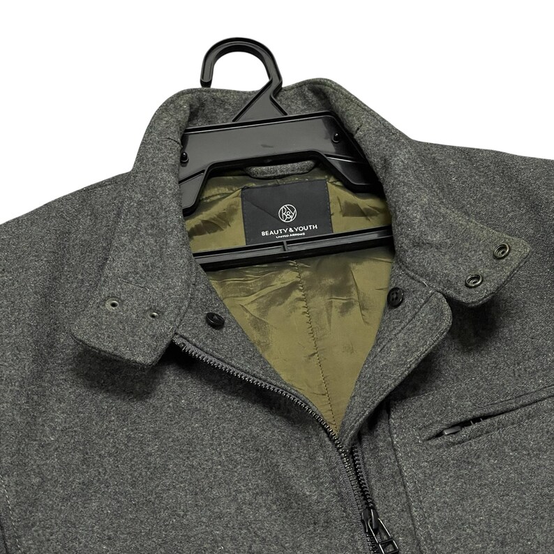 May include: A gray wool jacket with a black zipper and a small pocket on the right side. The jacket has a collar and is hanging on a black hanger. The interior lining is olive green, and the label reads "BEAUTY & YOUTH UNITED ARROWS".