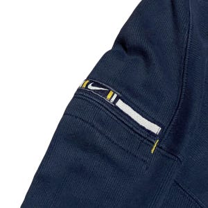 Puede incluir: Primer plano de una manga de sudadera azul marino. La manga presenta un parche rectangular con el logo de Nike en blanco, amarillo y negro. Las costuras son visibles y la tela tiene una textura acanalada.