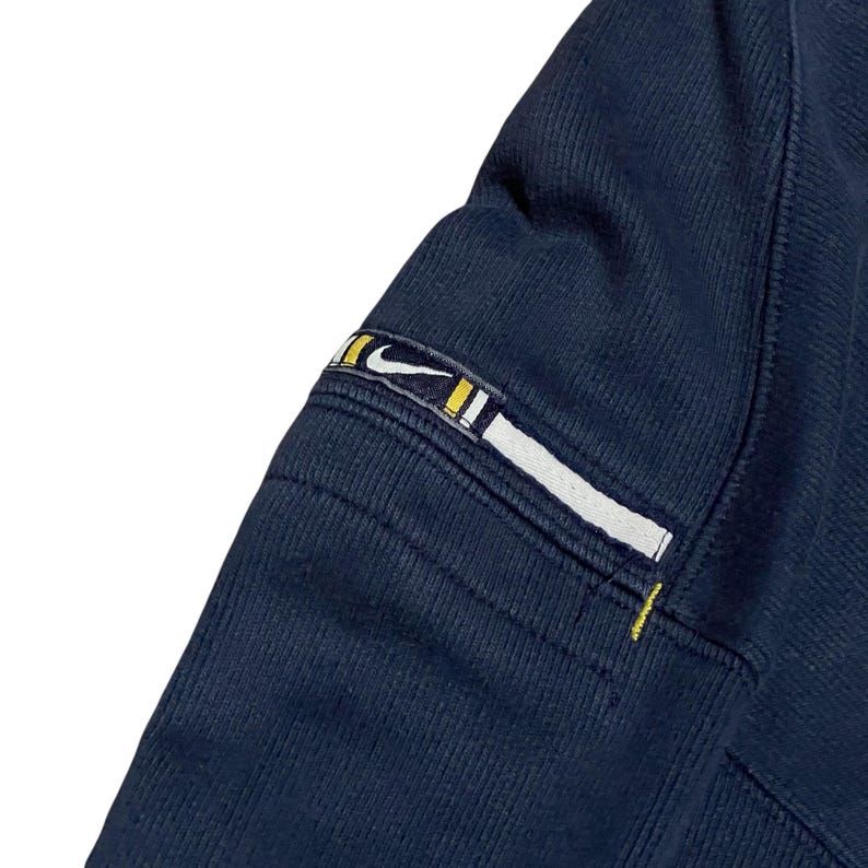 Puede incluir: Primer plano de una manga de sudadera azul marino. La manga presenta un parche rectangular con el logo de Nike en blanco, amarillo y negro. Las costuras son visibles y la tela tiene una textura acanalada.