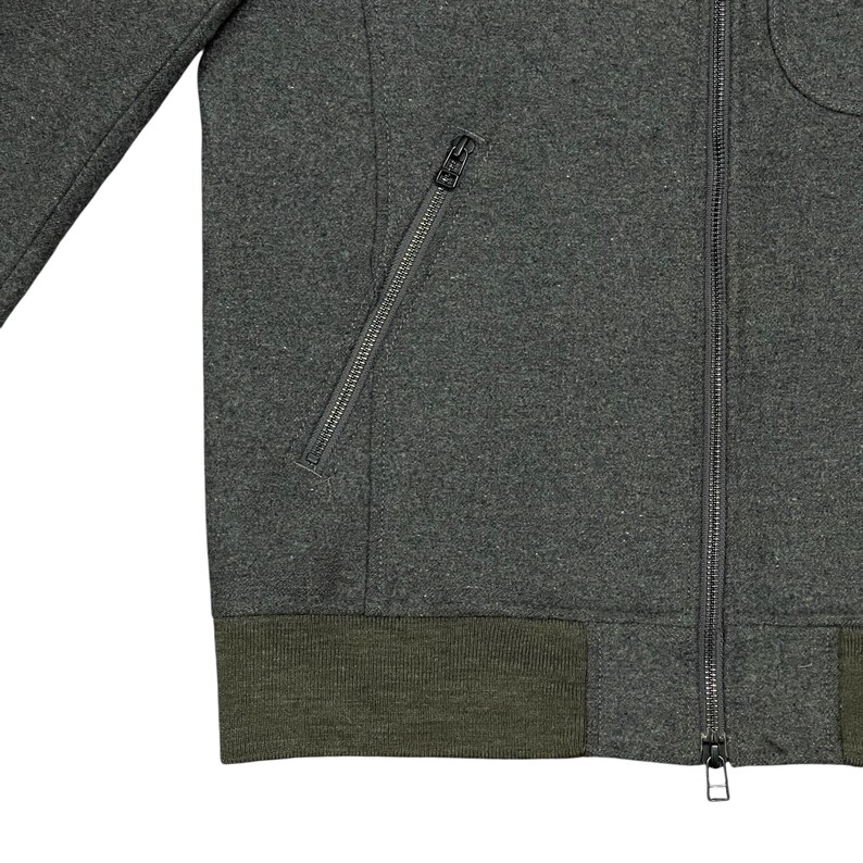 May include: Close-up of a dark gray zip-up jacket with a diagonal zippered pocket on the left side. The jacket has a dark green ribbed hem and cuffs. The zipper is silver.