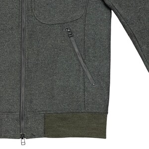 May include: A dark gray jacket with a full zipper closure and a diagonal zippered pocket. The jacket has a rounded pocket and a ribbed olive green hem. The jacket's fabric appears to be a wool blend.