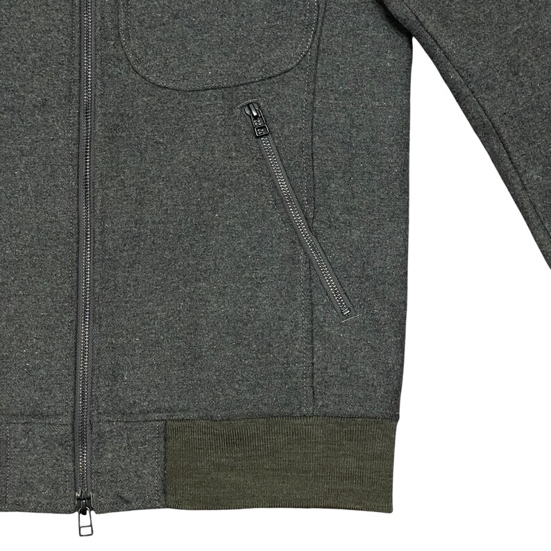 May include: A dark gray jacket with a full zipper closure and a diagonal zippered pocket. The jacket has a rounded pocket and a ribbed olive green hem. The jacket's fabric appears to be a wool blend.