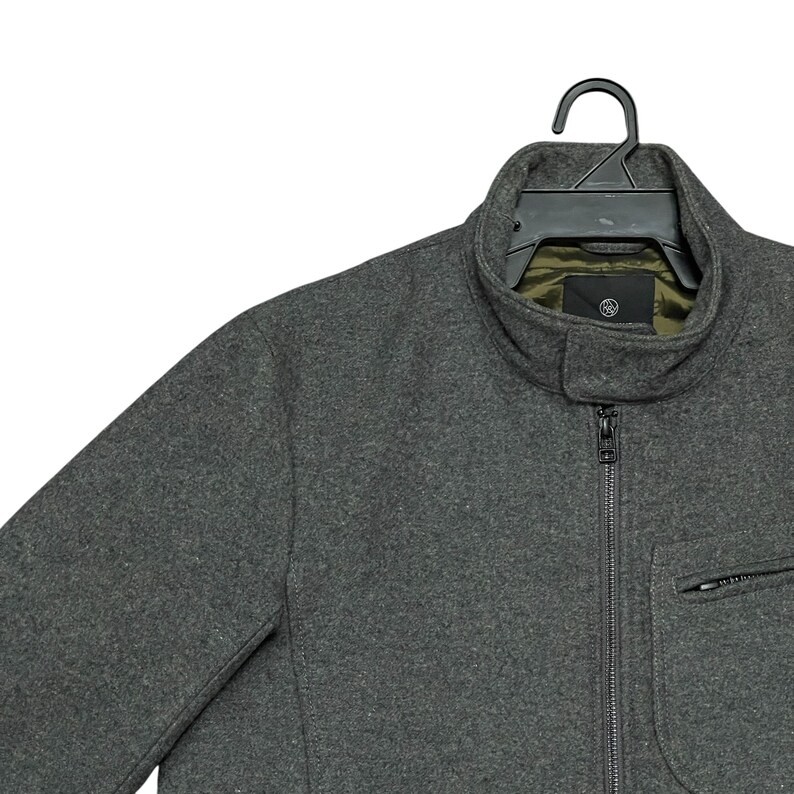 May include: A charcoal gray jacket with a full zipper closure and a small zippered pocket. The jacket has a high collar and is hanging on a black hanger. The interior lining is olive green.