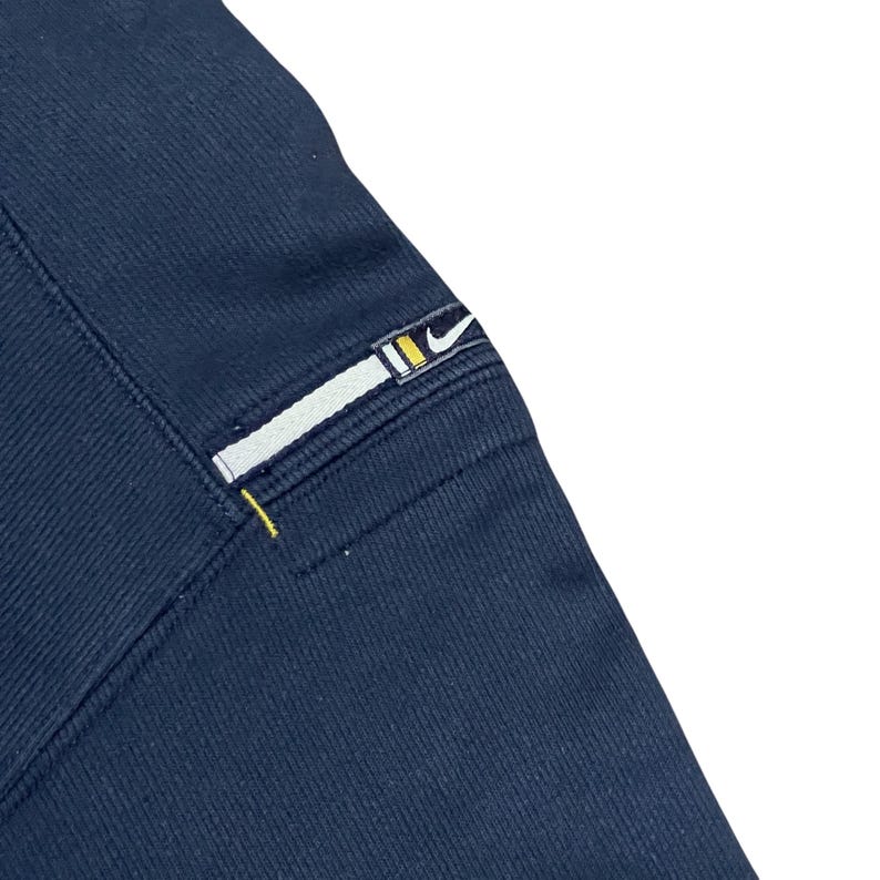 Puede incluir: Primer plano de tela azul marino con un parche rectangular. El parche presenta una franja blanca con un dise&ntilde;o amarillo y negro, y un peque&ntilde;o logotipo de Nike. Se ve una costura amarilla.