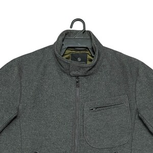 May include: A charcoal gray jacket with a high collar and a full-length zipper. The jacket has a single pocket on the chest and is hanging on a black hanger. The interior lining is olive green.
