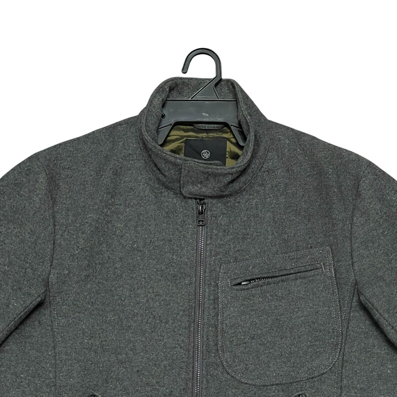 May include: A charcoal gray jacket with a high collar and a full-length zipper. The jacket has a single pocket on the chest and is hanging on a black hanger. The interior lining is olive green.