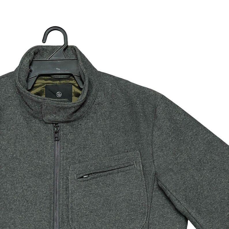 May include: A charcoal gray jacket with a high collar and a front zipper. The jacket has a single chest pocket with a zipper and is hanging on a black hanger. The interior lining is olive green.