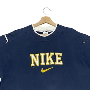 Puede incluir: Sudadera Nike azul marino con logo blanco y amarillo. La palabra "NIKE" est&aacute; bordada en letras amarillas con un swoosh amarillo debajo. El cuello es blanco con ribete azul marino.