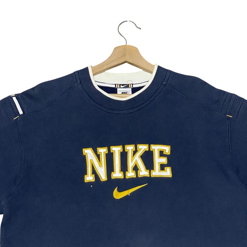 Puede incluir: Sudadera Nike azul marino con logo blanco y amarillo. La palabra "NIKE" est&aacute; bordada en letras amarillas con un swoosh amarillo debajo. El cuello es blanco con ribete azul marino.