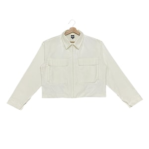 May include: Cream-colored cropped jacket with a collared neckline, two front pockets, and a full-length zipper. The jacket is hanging on a wooden hanger against a white background. The sleeves are long and the cuffs are buttoned.