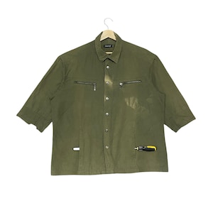 May include: Olive green button-down shirt with short sleeves. Features two chest pockets with zippers and a small tool detail on the lower right side. The shirt is hanging on a wooden hanger.