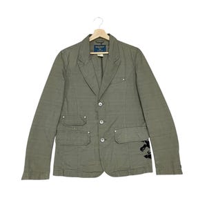 May include: An olive green blazer with a notched lapel, three-button fastening, and two front pockets. The jacket has a tailored fit and features decorative stitching and a small black graphic on the lower right side. It is displayed on a wooden hanger.