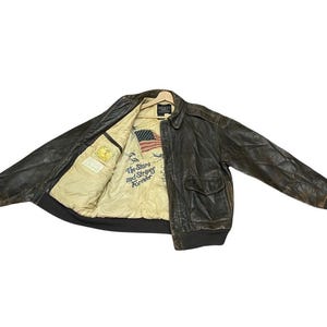 Könnte beinhalten: Braune Lederjacke im Bomber-Stil. Die Jacke hat zwei Vordertaschen sowie einen gerippten Saum und Bündchen. Das Innenfutter zeigt eine amerikanische Flagge und den Text "The Stars and Stripes Forever."