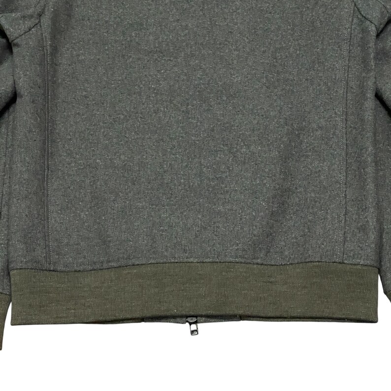 May include: A gray wool jacket with a dark olive green ribbed hem and cuffs. The jacket has a zipper closure. The fabric has a textured appearance.