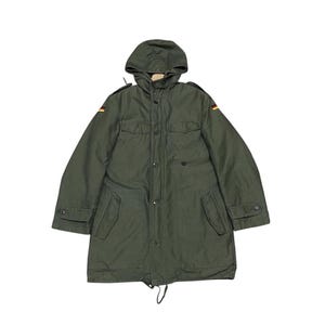 Könnte beinhalten: Olivgrüner Parka mit Kapuze, Knopfverschluss und Taschen. Die Jacke hat die deutsche Flagge auf den Schultern. Ein Kordelzug ist am Saum sichtbar. Klassischer Militärstil.
