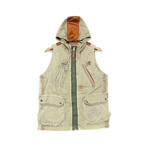 Puede incluir: Un chaleco sin mangas beige con capucha y capucha y detalles marrones. El chaleco tiene múltiples bolsillos, cierre de cremallera y una etiqueta con texto. El chaleco está colgado de una percha de madera.