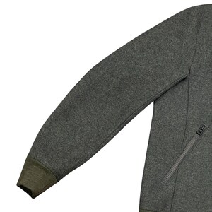 May include: A dark gray jacket with a zipper pocket and a dark green cuff. The jacket is made of a soft, textured material. The zipper is silver.