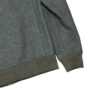 May include: A close-up of a gray jacket with a dark green ribbed hem and cuffs. The jacket has a zipper and a seam detail running down the sleeve. The fabric appears to be a wool blend.