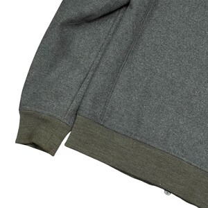 May include: Close-up of a gray sweatshirt with a dark green ribbed hem and cuffs. The sweatshirt has a simple design with a seam running down the sleeve. The fabric appears to be a soft, textured material.