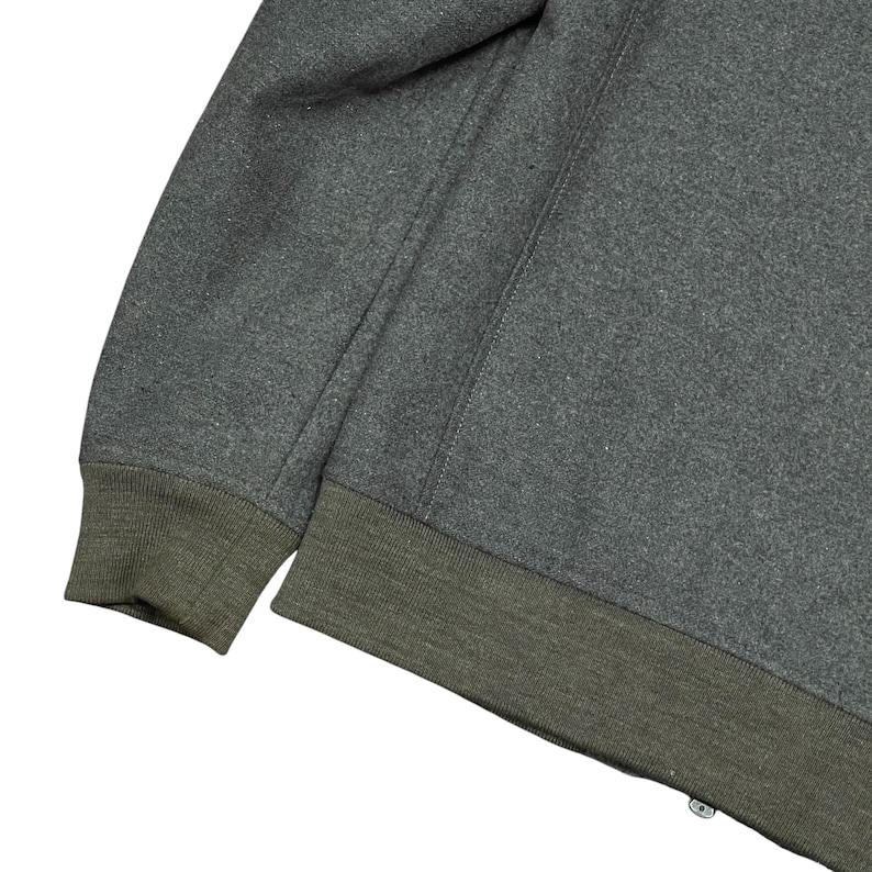 May include: Close-up of a gray sweatshirt with a dark green ribbed hem and cuffs. The sweatshirt has a simple design with a seam running down the sleeve. The fabric appears to be a soft, textured material.