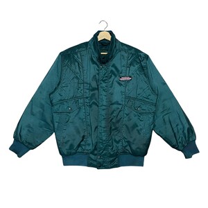 May include: A dark teal quilted jacket featuring a high collar, long sleeves with ribbed cuffs, and a ribbed hem. The front includes a central zipper closure with a snap-button placket and two front pockets. A Honda logo patch is on the chest.